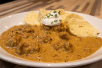 Bowl of Szegedin Goulash with creamy paprika sauce, sour cream, chives, and bread slices on a white plate.