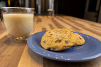 Two Chocolate Chip Cookies stacked on a blue plate next to a glass of milk on a wood table.