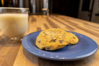Two Chocolate Chip Cookies on a blue plate next to a glass mug of milk on a wood counter.