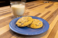 Two Chocolate Chip Cookies on a blue plate beside a glass of milk on a wood table.