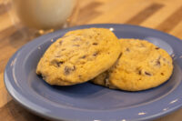 Two Chocolate Chip Cookies stacked on a blue plate with a glass of milk blurred behind them.