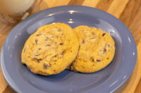Two golden Chocolate Chip Cookies on a blue plate with a glass of milk behind them.