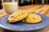 Two Chocolate Chip Cookies on a blue plate with a glass of milk behind them on a wooden table.