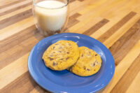Two Chocolate Chip Cookies on a blue plate with a glass of milk behind them on a wood table