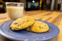 Two Chocolate Chip Cookies stacked on a blue plate with a glass of milk on a wooden table.