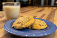 Two Chocolate Chip Cookies stacked on a blue plate with a glass of milk behind them on a wood table.