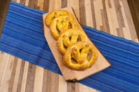 Three golden Czech Soft Pretzel Preclík with coarse salt on a wooden board over a blue striped runner.