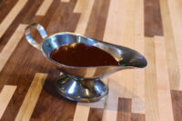 Silver gravy boat filled with dark brown Demi-Glace Sauce on a striped wooden surface.
