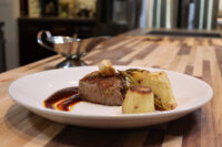 Seared filet mignon with demi-glace sauce, roasted garlic, rosemary, and potato wedges on a white plate.