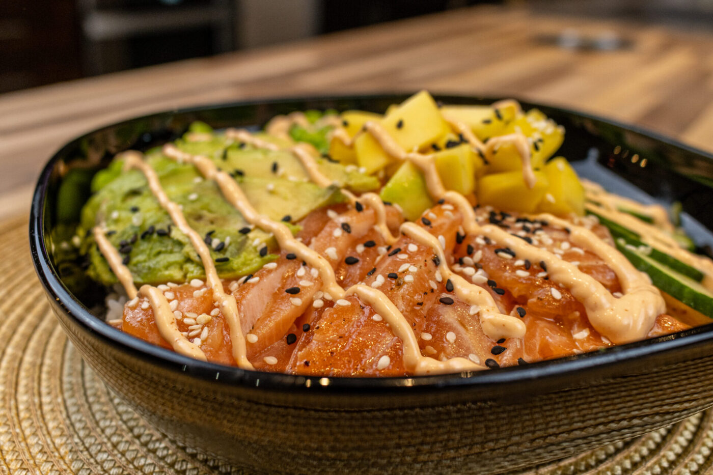 Black bowl of Poke Bowl with Salmon topped with avocado, mango, sesame seeds and creamy drizzle.