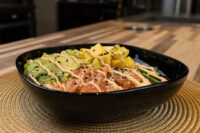 Black bowl of Poke Bowl with Salmon topped with avocado, mango, cucumber, and spicy mayo drizzle.