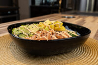 Black bowl of Poke Bowl with Salmon topped with mango, avocado, and spicy mayo drizzle.