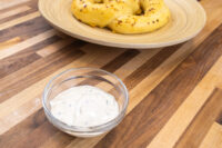 Glass bowl of Garlic Dipping Sauce for Pretzels with visible chive flecks, pretzel rolls on a wooden plate behind it