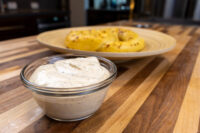 Glass bowl of Mustard Dipping Sauce for Soft Pretzels on a wood counter with two pretzels behind it.