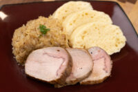 Sliced houskový knedlík fanned beside sauerkraut and pork loin on a dark red plate.