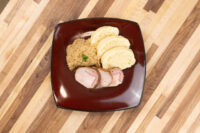 Sliced houskový knedlík rounds beside pork loin and sauerkraut on a dark square plate