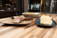 Sliced roast pork, a bowl of stewed sauerkraut, and a sliced knedlík for Vepřo-knedlo-zelo on a wooden counter.