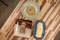Sliced houskový knedlík on a dark plate beside a bowl of rice and a rolled meat dish.
