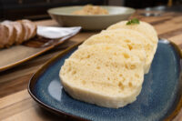 Sliced houskový knedlík on a blue plate, with bread and a bowl blurred in the background
