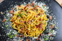 Twirled Spaghetti Carbonara on a black plate topped with crispy bacon bits, herbs and grated cheese.
