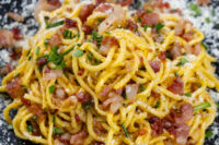 Spaghetti Carbonara tossed with crispy bacon, herbs, and cheese on a dark speckled plate.