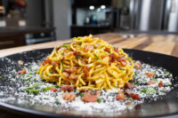 Golden spaghetti carbonara with crispy bacon bits, grated cheese, and herbs on a black plate
