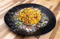 Plate of Spaghetti Carbonara with bacon bits, herbs, and grated cheese on a black dish.