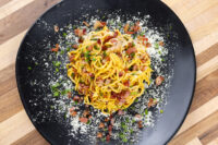 Overhead view of Spaghetti Carbonara with crisp bacon bits, herbs, and grated cheese on a black plate.