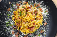 Overhead shot of Spaghetti Carbonara topped with crispy bacon, herbs, and grated cheese on a black plate.