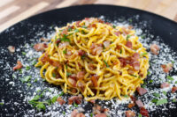 Twirled Spaghetti Carbonara with bacon bits, herbs, and grated cheese on a black plate.
