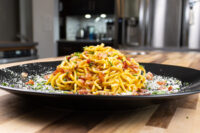 Mounded Spaghetti Carbonara with bacon bits, herbs, and grated cheese on a black plate.