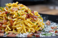 Close-up of Spaghetti Carbonara piled high with crispy bacon, chives, and grated cheese on a dark plate.