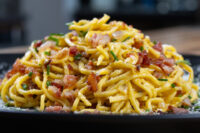 A mound of Spaghetti Carbonara with crispy bacon, chives, and grated cheese on a dark plate.