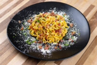 Plate of Spaghetti Carbonara with crispy bacon bits, grated cheese, and herbs on black plate.