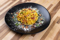 Black plate of Spaghetti Carbonara with bacon bits, herbs, and grated cheese on a wooden table