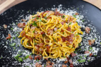 Plate of Spaghetti Carbonara with bacon bits, herbs, and grated cheese on a black dish.
