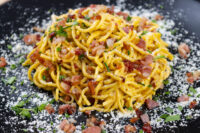 Golden Spaghetti Carbonara twirled on a black plate with crispy bacon bits, herbs, and grated cheese.
