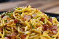 Close-up of Spaghetti Carbonara with crispy bacon bits, chives, and grated cheese on a dark plate