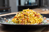 Twirled Spaghetti Carbonara topped with crispy bacon, herbs, and grated cheese on a black plate.