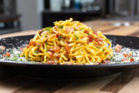 Heaping plate of Spaghetti Carbonara with crispy bacon, herbs, and grated cheese on black plate.