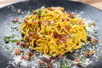 Golden-sauced Spaghetti Carbonara topped with bacon bits, herbs, and grated cheese on a black plate.