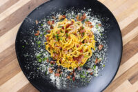 Spaghetti Carbonara twirled on a black plate, coated in yellow sauce with crispy bacon, cheese, and herbs