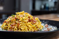 Heaping plate of Spaghetti Carbonara with crispy bacon, chives, and grated cheese on black dish.