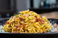 Golden Spaghetti Carbonara piled high with crispy bacon, chives, and grated cheese on a black plate.