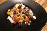 Black plate of Greek Salad with diced tomatoes, peppers, red onion, olives, and crumbled feta.