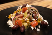 Greek Salad with feta, kalamata olives, tomatoes, red onion and peppers on a black plate.