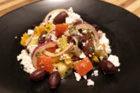 Black plate of Greek Salad with tomato, pepper, red onion, olives and crumbled feta.