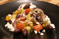 Greek Salad with diced peppers, tomato, red onion, kalamata olives and crumbled feta on a black plate.