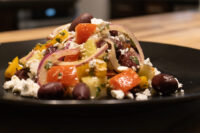 Black plate of Greek Salad with diced tomatoes, red onion, kalamata olives, and feta on a wooden table.