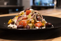 Greek Salad on a black plate with roasted vegetables, red onion, olives, and crumbled feta.